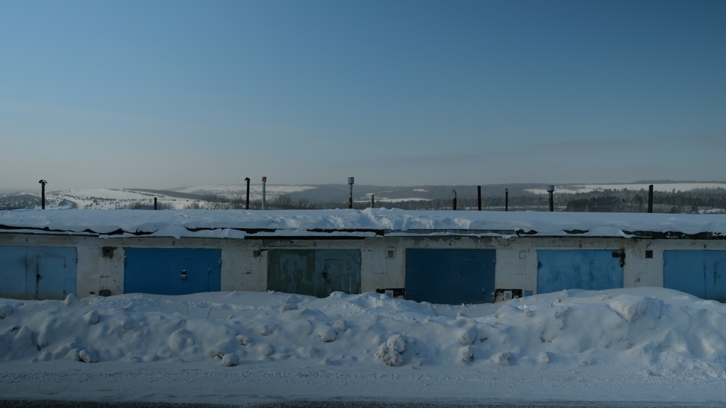 Photo of one-story storage units that have been snowed in due to plowing; a rural, snowy landscape rises above to a blue sky.